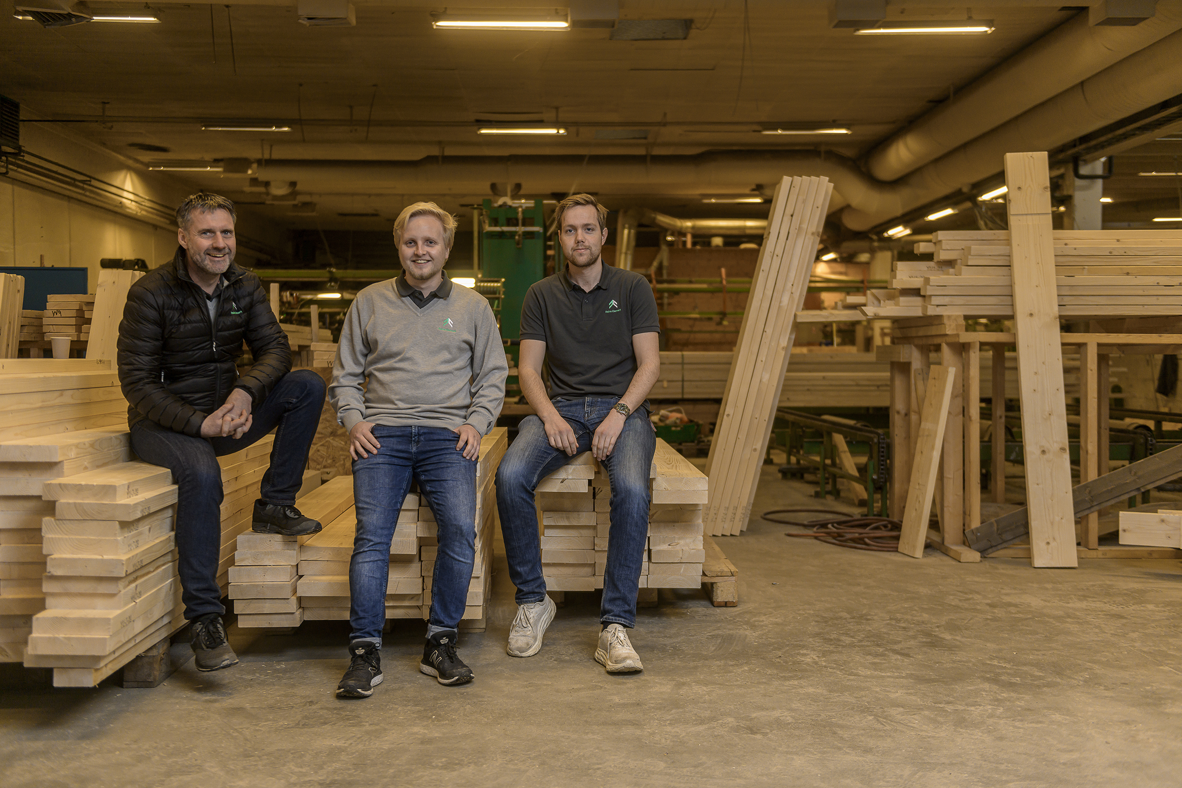Tre ansatte sitter på stabler av precut materiale og smiler mot kamera i fabrikken til Reine Element